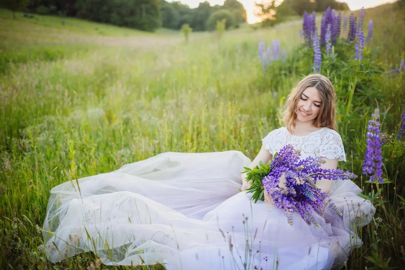 Eine junge Frau in prächtigem Kleid sitzt mit einem Strauß violetter Blumen auf einem grünen Feld.